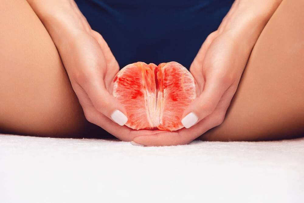 A woman holds half a grapefruit between her legs with two hands